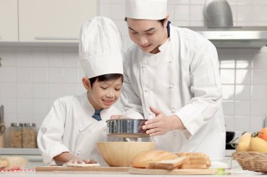 Mutfakta şapka pişiren, şef üniformalı mutlu Asyalı oğul ve baba. Tatlı çocuk babasının un karıştırmasına yardım ediyor, yoğurmadan önce ekmek hamuru hazırlıyor, ebeveyn ve çocuk fırın yapıyor..