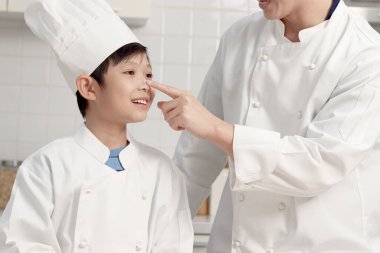 Mutfakta şapkalı mutlu Asyalı oğul ve baba, ebeveyn ve çocuk ekmek hamuru hazırlıyor, baba oğluna burun buruna dokunuyor, şirin aile fırın yaparken birlikte eğleniyor..