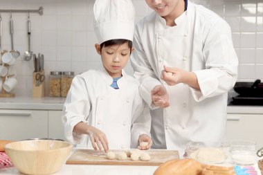 Mutlu Asyalı oğul ve aşçı üniformalı baba mutfakta birlikte yemek pişiriyorlar baba ve çocuk ekmek hamuru hazırlıyor ve yoğuruyor şirin bir aile fırın yapıyor. Çocuk öğrenci yemek yapmayı öğreniyor..