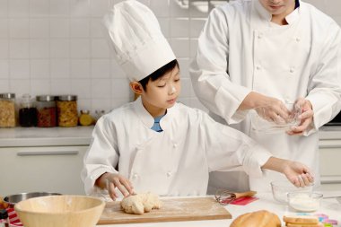 Mutlu Asyalı oğul ve aşçı üniformalı baba mutfakta birlikte yemek pişiriyorlar baba ve çocuk ekmek hamuru hazırlıyor ve yoğuruyor şirin bir aile fırın yapıyor. Çocuk öğrenci yemek yapmayı öğreniyor..