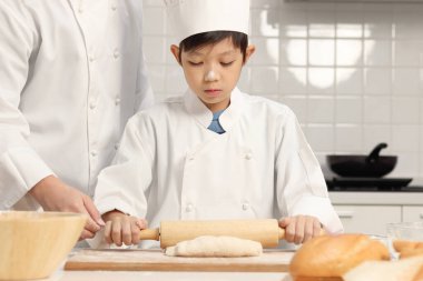 Mutlu Asyalı oğul ve aşçı üniformalı baba, mutfakta birlikte yemek pişiren şapkalı yakışıklı çocuk merdaneyle hamuru yoğuruyor, aile içinde fırın yapan ebeveyn ve çocuk. Çocuk öğrenci ekmek pişirmeyi öğreniyor..