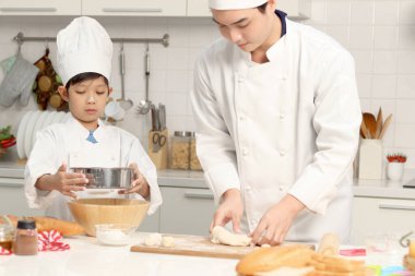 Mutfakta şapka pişiren, şef üniformalı mutlu Asyalı oğul ve baba. Tatlı çocuk babasının un karıştırmasına yardım ediyor, yoğurmadan önce ekmek hamuru hazırlıyor, ebeveyn ve çocuk fırın yapıyor..