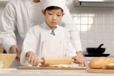 Mutlu Asyalı oğul ve baba aşçı üniforması içinde mutfakta şapkalı yemek pişiriyor, tatlı çocuk hamuru oklavayla yoğuruyor, baba çocuğa ekmek pişirmede yardım ediyor ve öğretiyor, harika bir aile birlikte fırın yapıyorlar..