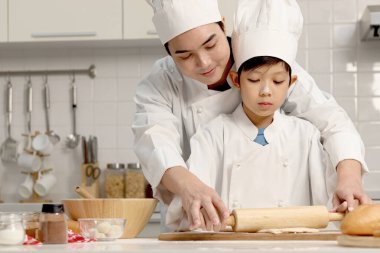 Mutlu Asyalı oğul ve baba aşçı üniforması içinde mutfakta şapkalı yemek pişiriyor, tatlı çocuk hamuru oklavayla yoğuruyor, baba çocuğa ekmek pişirmede yardım ediyor ve öğretiyor, harika bir aile birlikte fırın yapıyorlar..