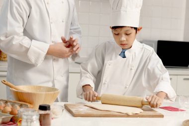 Mutlu Asyalı oğul ve baba aşçı üniforması içinde mutfakta şapkalı yemek pişiriyor, tatlı çocuk hamuru oklavayla yoğuruyor, baba çocuğa ekmek pişirmede yardım ediyor ve öğretiyor, harika bir aile birlikte fırın yapıyorlar..
