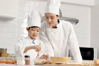 Mutlu Asyalı oğul ve aşçı üniformalı baba mutfakta birlikte yemek pişiriyorlar baba ve çocuk ekmek hamuru karıştırıp yoğuruyorlar sevimli bir aile fırını yapıyorlar. Çocuk öğrenci ekmek pişirmeyi öğreniyor.