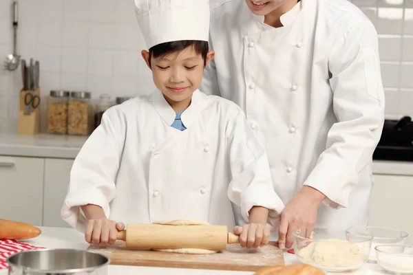 快乐的亚洲儿子和父亲 身穿厨师制服 戴着帽子一起在厨房做饭 可爱的男孩用滚动的别针揉搓面团 父母和孩子在家里做糕点 孩子们学习烤面包 — 图库照片