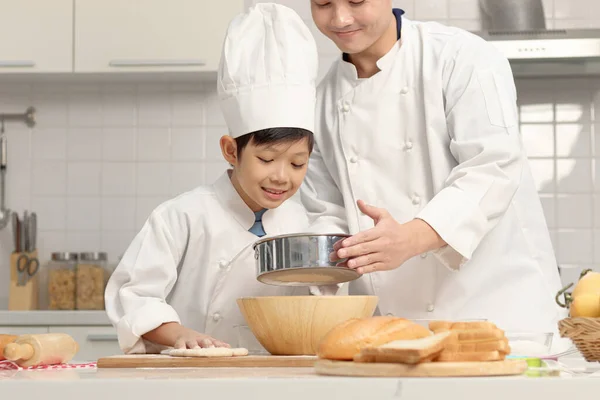 快乐的亚洲儿子和父亲 穿着厨师制服 戴着帽子 在厨房做饭 可爱的男孩帮助爸爸把面粉筛入碗中 在揉碎之前准备好面包面团 父母和家里的孩子一起做糕点 — 图库照片