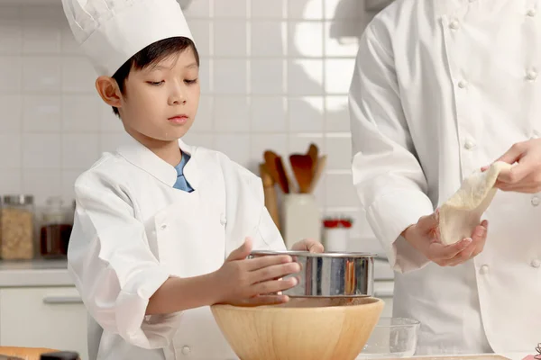快乐的亚洲儿子和父亲 穿着厨师制服 戴着帽子 在厨房做饭 可爱的男孩帮助爸爸把面粉筛入碗中 在揉碎之前准备好面包面团 父母和家里的孩子一起做糕点 — 图库照片