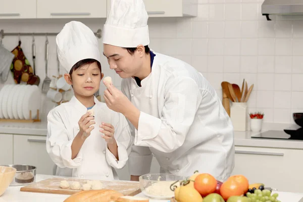 快乐的亚洲儿子和父亲 身穿厨师制服 戴着帽子在厨房里 父亲和孩子们一起揉搓和准备面包面团 可爱的家庭一起做糕点 小学生学习烤面包 — 图库照片
