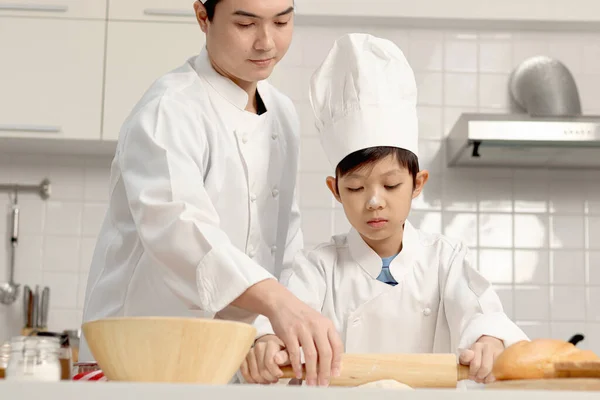 快乐的亚洲儿子和父亲 身穿厨师制服 戴着帽子一起在厨房做饭 可爱的男孩用滚动的别针揉搓面团 父母和孩子在家里做糕点 孩子们学习烤面包 — 图库照片