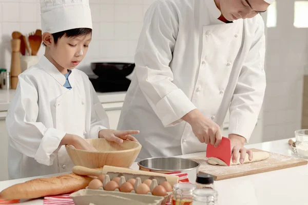 快乐的亚洲儿子和父亲 身穿厨师制服 头戴帽子 一起在厨房做饭 父亲和儿子一起搅拌和揉搓面包面团 可爱的家庭做糕点 小学生学习烤面包 — 图库照片