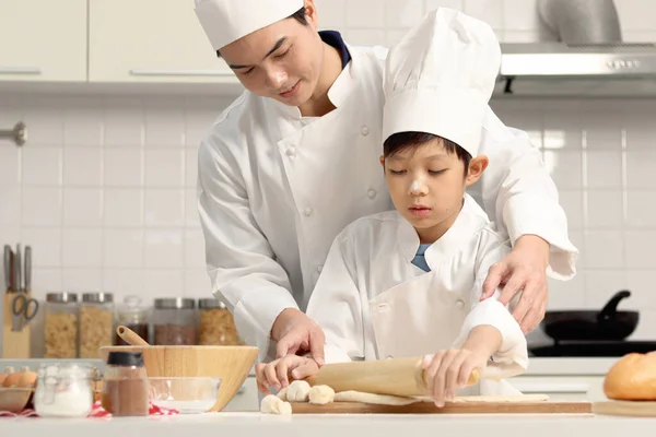 快乐的亚洲儿子和身穿厨师制服的父亲在厨房里用帽子做饭 可爱的男孩用滚动的别针揉面 爸爸帮助和教孩子烘焙面包 可爱的家庭一起做糕点 — 图库照片