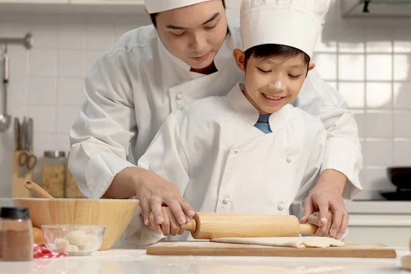 快乐的亚洲儿子和身穿厨师制服的父亲在厨房里用帽子做饭 可爱的男孩用滚动的别针揉面 爸爸帮助和教孩子烘焙面包 可爱的家庭一起做糕点 — 图库照片