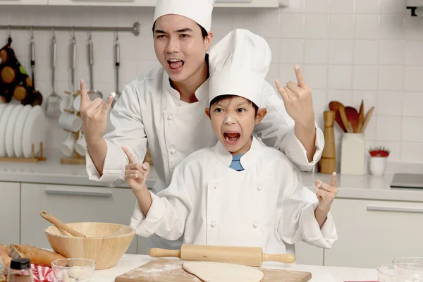 Happy Asian son kid and father in chef uniform with hat showing rock and roll finger gesture at kitchen together, dad parent and boy child having fun during baking bread, cute family making bakery.