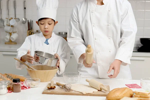 Happy Asian son and father in chef uniform with hat cooking at kitchen. cute boy child helps dad sifting flour into bowl, preparing bread dough before kneading, parent and kid in family making bakery.