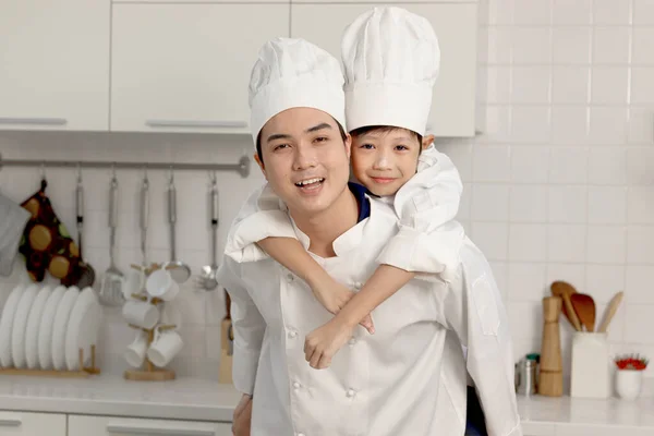 快乐的亚洲儿子和父亲 身穿厨师制服 戴着帽子在厨房里 爸爸骑着他的孩子 父母和男孩一起玩耍 在烘焙面包的过程中一起玩乐 可爱的家庭一起做糕点 — 图库照片