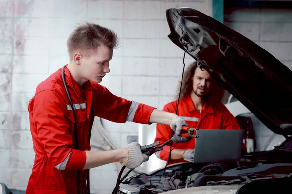 身穿红色制服的机械师通过跳线给汽车蓄电池充电 同事在车库里一起工作 技师团队在维修车间修理客户的汽车 — 图库照片