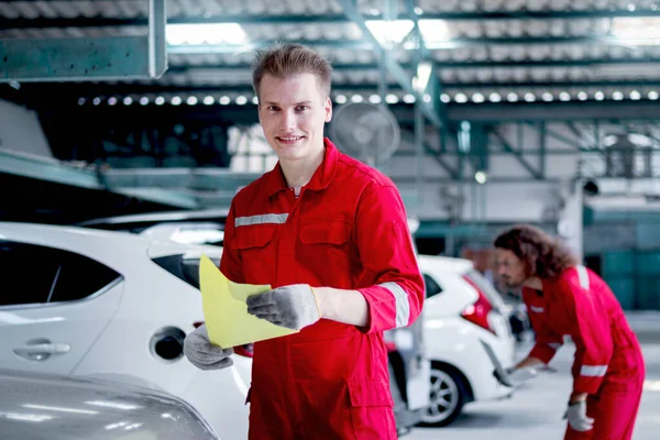 穿着红色制服 面带微笑的机械师的画像 站在车库里的汽车旁 汽车技师在维修车间检查维修客户的汽车 — 图库照片
