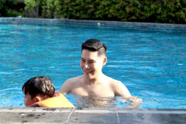 Mutlu Asyalı bir baba ve mavi su havuzunda can yeleği olan küçük bir oğlan çocuğu, baba çocuğa yüzmeyi öğretiyor, çocuk yüzmeyi öğreniyor, aile yüzücüsünü rahatlatıyor yazın birlikte eğlenceli aktiviteler yapıyorlar.. 