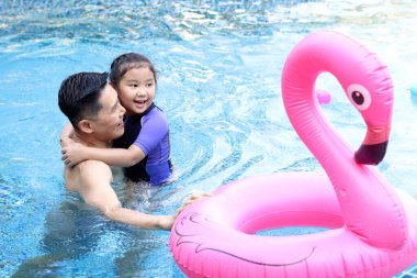 Mavi su havuzunda pembe flamingo plastik oyuncaklı mutlu Asyalı bir baba ve küçük bir kız çocuğu, baba çocuğa yüzmeyi öğretiyor, aile yüzücüsünü rahatlatıyor yazın birlikte eğlenceli aktiviteler yapıyorlar..