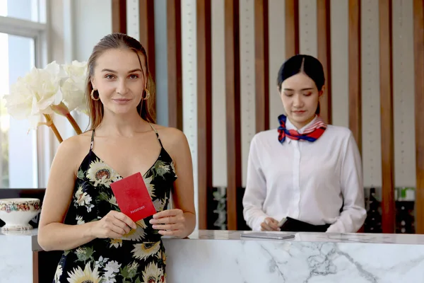 Happy beautiful tourist woman customer showing passport during checking in at hotel reception counter desk with Asian female receptionist, female guest checks in at hotel on holiday vacation.