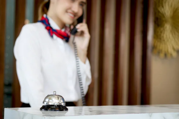 Silver bell on hotel reception service desk with blurred background of smiling Asian female receptionist talks to customer guest on telephone at reception counter desk hotel, booking hotel on vacation
