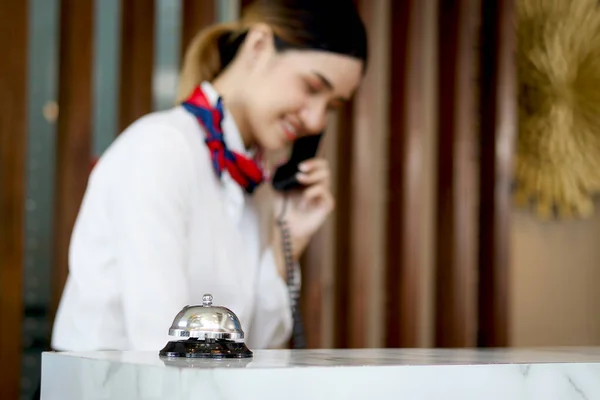 Silver bell on hotel reception service desk with blurred background of smiling Asian female receptionist talks to customer guest on telephone at reception counter desk hotel, booking hotel on vacation