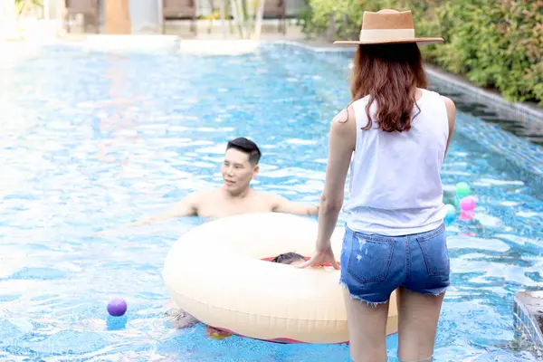 Back view of beautiful young Asian woman in summer hat has a great time at pool party, cheerful female and friend have fun with summer outdoor activity, happy girl relaxing by the pool.