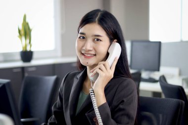 Güzel Asyalı kadın operatör ofis masasında çalışan meslektaşları ile sabit telefon kullanarak müşteriyle konuşuyor. İş yerinde gülümseyen kadın resepsiyonist