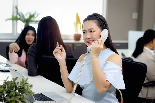 Beautiful Asian woman officer talking with customer by using landline phone at working desk in office with blurred background of busy working colleagues. Attractive smiling female worker at workplace.