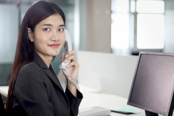 Beautiful Asian woman operator officer talking with customer by using landline phone at working desk in office with busy working colleagues blurred background. Smiling female receptionist at workplace