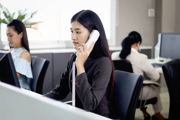 Beautiful Asian woman operator officer talking with customer by using landline phone at working desk in office with busy working colleagues blurred background. Smiling female receptionist at workplace