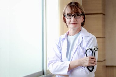 Gözlüklü, steteskop tutan, hastane hastanesinde kolları çapraz duran, beyaz iş elbiseli, kendine güvenen kadın doktor portresi. Profesyonel kıdemli kadın doktor.. 