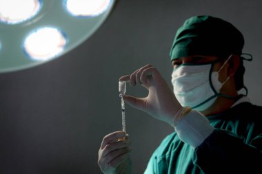 Profesyonel cerrah elinde şırınga tutuyor, parlak ışığın altında duruyor, ameliyat için cerrahi ekipman kullanıyor. Cerrahi ekip ameliyat hastası, sağlık sigortası