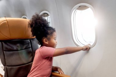 Mutlu kıvırcık saçlı Afrikalı kız çocuk yolcu uçağın penceresinin yanındaki rahat koltukta oturuyor, çocuk pencereye dokunmak için elini uzatıyor, tatil seyahati ve macera gezisi.