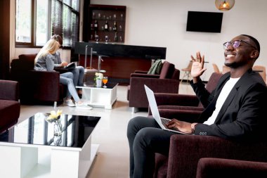Havaalanı iş yeri lobisindeki koltukta otururken dizüstü bilgisayarda çalışan Afrikalı iş adamı mutlu bir şekilde gülümseyen erkek online toplantı yapıyor ve selamlaşmak için el sallıyor..