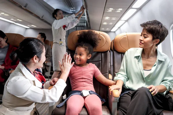Friendly air hostess take care and help kid passenger on airplane, cheerful flight attendant and curly hair African girl giving high five on aircraft, child traveling by plane, airline transportation