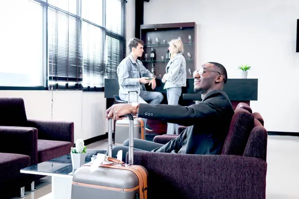 African  businessman with luggage sitting on sofa in airport business lounge, happy smiling male passenger sitting and resting at waiting area of airport, waiting flight at airport departure lounge