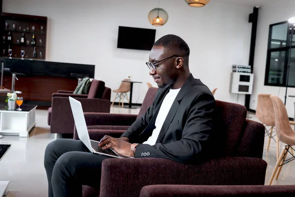African businessman working on laptop computer while sitting on sofa in airport business lounge or hotel lobby, happy smiling male has online conference meeting during waiting flight at airport lounge