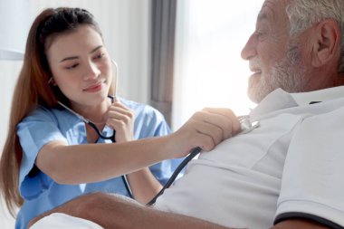 Steteskop kullanarak gülümseyen güzel bayan doktor hastane yatağında yatan yaşlı hasta kalbini dinle. Hemşire kendini hasta hissediyor ve evde dinleniyor. Sağlık hizmetleri..