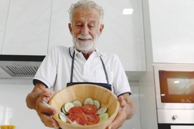Yaşlı adam mutfak tezgahında elinde taze sebze kasesi, kıdemli adam evde organik malzemelerle sağlıklı yiyecekler hazırlıyor ve pişiriyor, olgun büyükbaba mutfakta yemek yapıyor..