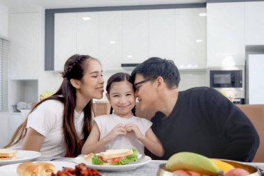 Mutlu aile yemek odasında yemek yer. Anne, baba, kız, yemek masasında oturur, kahvaltı sırasında eğlenir, kalbi elle şekillendiren ebeveyn, öpen kız çocuğu. Aile birlikte yemek yemekten hoşlanır.