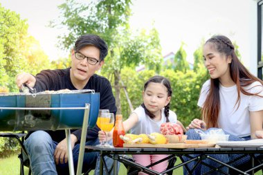 Neşeli ebeveyn ve çocuk piknik partisinde barbekü yapıyor, yaz yeşil bahçesinde birlikte eğleniyorlar, baba ve kız arka bahçede yiyip içmenin keyfini çıkarıyorlar, mutlu aile bağları.