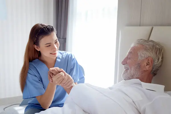 Smiling beautiful caring female doctor holding hand of male senior patient who lying in hospital bed. Nurse takes care elderly man feeling sick need to rest in bed at home, medical elderly health care