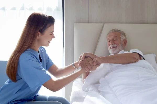 Smiling beautiful caring female doctor holding hand of male senior patient who lying in hospital bed. Nurse takes care elderly man feeling sick need to rest in bed at home, medical elderly health care