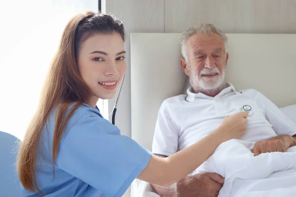Smiling beautiful caring female doctor using stethoscope listen to senior patient heart who lying in hospital bed. Nurse takes care man feel sick and rest in bed at home, medical elderly health care.
