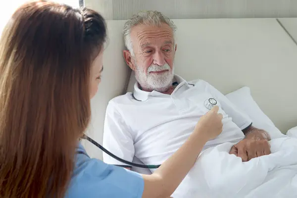 Elderly man feels sick and rest in bed at home with caring female nurse or doctor using stethoscope listen to patient heart, nursing senior patient at hospital or house, medical elderly health care.