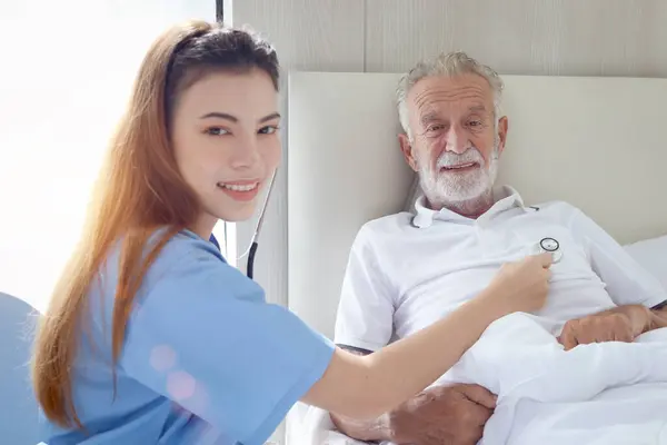Elderly man feels sick and rest in bed at home with caring female nurse or doctor using stethoscope listen to patient heart, nursing senior patient at hospital or house, medical elderly health care.