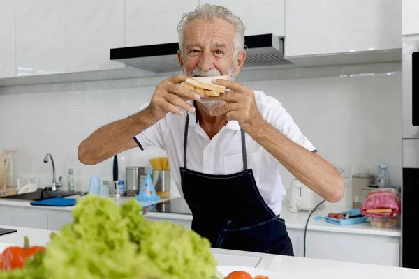 Happy smiling elderly man wearing apron, standing at kitchen counter. Senior man holding bread sandwich and ready to eat it, mature grandfather enjoys cooking meal by himself and eating at home.
